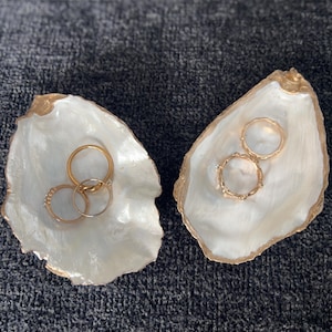 May include: Two white oyster shell ring dishes with gold accents. The dishes are filled with gold rings.