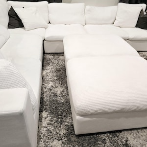 May include: A large, white sectional sofa with a matching ottoman. The sofa has multiple sections and is adorned with white pillows and a dark gray pillow. The ottoman is rectangular and covered in the same textured fabric as the sofa.