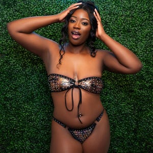 Puede incluir: Una mujer lleva un traje de baño de dos piezas negro y dorado brillante con un lazo en la parte delantera. Está de pie frente a una pared verde con un letrero de neón que dice "Amelia Monae LLC".