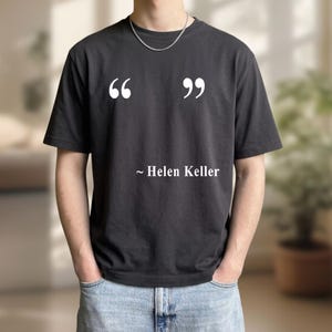 May include: Black t-shirt with white quotation marks and the text "~ Helen Keller." The shirt is a dark charcoal color and has short sleeves. The person is wearing blue jeans and a silver chain.