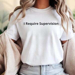 May include: White t-shirt with the phrase "I Require Supervision" printed in black. The shirt is a crew neck style and appears to be made of a soft material. The model is wearing a beige jacket and blue jeans.