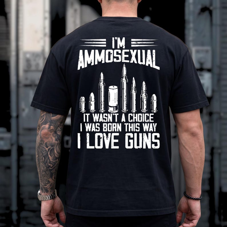 Puede incluir: Camiseta negra con texto blanco e ilustraciones de balas. El texto dice "I'M AMMOSEXUAL, IT WASN'T A CHOICE I WAS BORN THIS WAY I LOVE GUNS". La camiseta tiene cuello redondo y mangas cortas.