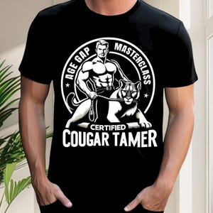 May include: Black t-shirt with a white graphic. The graphic features a muscular man holding a rope attached to a cougar. Text on the shirt reads "AGE GAP MASTERCLASS CERTIFIED COUGAR TAMER".