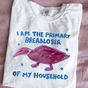 May include: A white short-sleeve t-shirt featuring a graphic of a pink duck. The text reads "I AM THE PRIMARY BREADLOSER OF MY HOUSEHOLD" in blue letters. Small blue stars surround the duck, and the shirt has a Comfort Colors brand label.