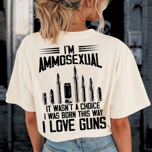 Puede incluir: Camiseta color crema con el texto "I'M AMMOSEXUAL" sobre una fila de ilustraciones de balas. Debajo, la camiseta dice "IT WASN'T A CHOICE I WAS BORN THIS WAY I LOVE GUNS."