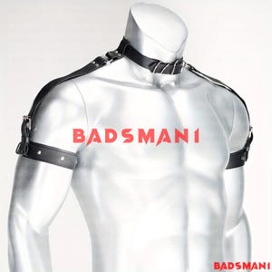 May include: Black leather harness with a choker-style collar and armbands. The harness features silver-tone hardware and adjustable straps. The word "BADSMAN1" is printed in red across the chest.