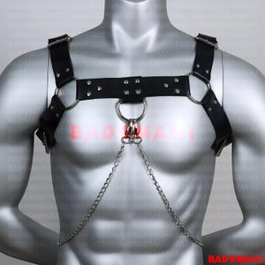 May include: Black leather chest harness with silver-tone metal rings and chain accents. The harness features adjustable straps and decorative rivets. The design includes a central ring with a dangling chain.