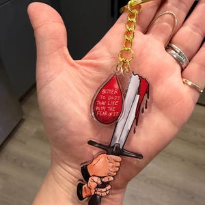 Puede incluir: Un llavero acrílico transparente con una cadena y un anillo dorados. El llavero presenta una espada con sangre roja goteando y una forma de lágrima roja con el texto "Better to do it than live with the fear of it."