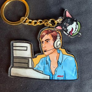 May include: An acrylic keychain featuring a cartoon illustration of a person with brown hair wearing a blue shirt and a headset while sitting at a computer. A small charm of a black and white dog with a green collar is attached to the gold metal ring.