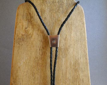 May include: A black braided bolo tie with a silver-toned tip. The tie is displayed on a wooden stand.