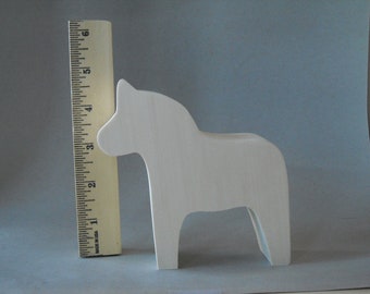 Puede incluir: Una figura de caballo Dala de madera blanca, de aproximadamente 10 cm de altura, está junto a una regla. El caballo tiene un diseño simple y estilizado, con un cuerpo curvo y una cabeza pequeña. La regla está marcada en pulgadas.