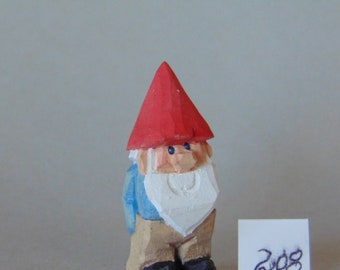 May include: A small wooden gnome figurine with a red hat, blue shirt, and white beard. The gnome is standing on a white surface.