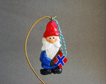 May include: A wooden Christmas ornament depicting a gnome wearing a red hat, blue coat, and white beard. The gnome is holding a small Norwegian flag.