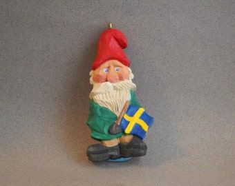 May include: A wooden gnome ornament with a red hat, white beard, and green clothing. The gnome is holding a small yellow and blue flag.