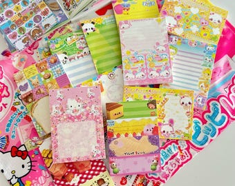 May include: A collection of colorful stationery items, including notepads and stickers, featuring cartoon characters and food illustrations. The items are in various shapes and sizes, with pink, yellow, and green dominating the color palette. Some items have Japanese text.