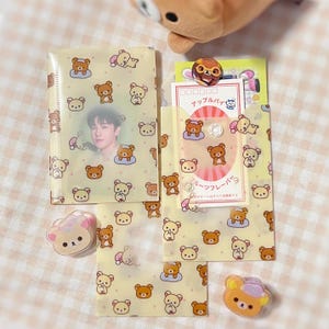 May include: A collection of stationery items with a repeating pattern of cartoon bears. Includes a clear plastic folder with a photo, a small pouch with cards, and bear-shaped accessories. The items are on a light yellow background.