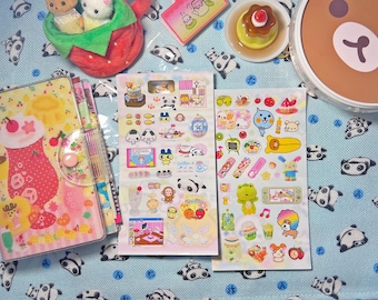 May include: A collection of colorful stickers, a planner, and plush toys are arranged on a light blue patterned surface. The stickers feature various cartoon characters and food items. A dessert and a container with a bear face are also visible.