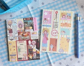 May include: Two illustrated notebooks with Japanese text and a blue pen on a blue and white patterned surface. The notebooks feature cartoon characters, food illustrations, and text. One notebook has a cafe theme, and the other has a summer theme.