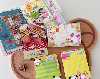 May include: A collection of colorful stationery items, including notebooks, sticky notes, and a small decorative figure. The items feature cute cartoon characters and food-themed illustrations, with a brown bear-shaped coaster in the background.