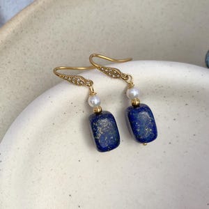 May include: A pair of gold-toned earrings featuring rectangular, deep blue lapis lazuli stones. Each earring has a small white pearl and a decorative top with small clear crystals. The earrings are displayed on a white, textured surface.