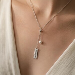 May include: A silver necklace with a delicate design. The necklace features a small pearl and a rectangular pendant with an engraved floral design. The necklace is worn against a white top.