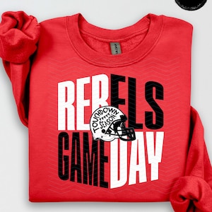 Może przedstawiać: Czerwona bluza z napisem "REBELS GAME DAY" w białych i czarnych literach. Grafika kasku do futbolu amerykańskiego znajduje się pomiędzy słowami. Na kasku napis "TOUCHDOWN SEASON".