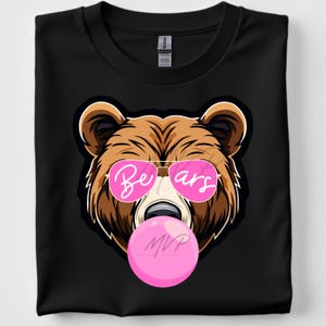 May include: Black t-shirt featuring a brown bear graphic wearing pink sunglasses with the word "Bears" and blowing a pink bubble. The bear graphic is outlined in black. The shirt is folded.