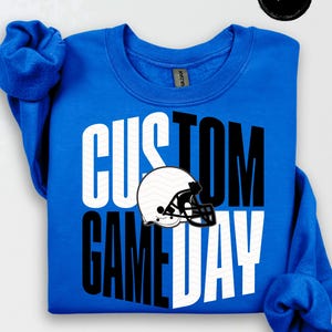 May include: A royal blue sweatshirt featuring the text "CUSTOM GAME DAY" in white and black. A white and black American football helmet graphic is positioned in the center. The brand logo is in the upper right corner.