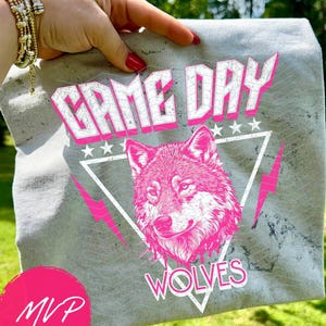 May include: Gray t-shirt with a pink wolf graphic, the words "GAME DAY" and "WOLVES" in white and pink, and pink lightning bolts. The shirt is held up by a hand with red nail polish and gold bracelets.