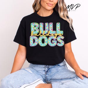 May include: Black t-shirt with the words "BULLDOGS" in large, colorful letters. The letters are outlined in green and filled with a floral pattern. The word "bulldogs" is in yellow script. The shirt is worn by a person.