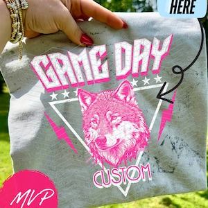 May include: Grey t-shirt featuring a pink wolf graphic and the words "GAME DAY CUSTOM". The design includes a triangle, stars, and lightning bolts. The shirt is held up by a hand with gold bracelets.