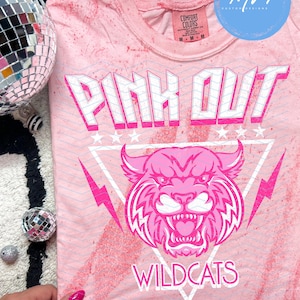 May include: Pink t-shirt with the words "PINK OUT" in white and pink block letters. Below the text is a pink wildcat graphic inside a white triangle. The shirt has a tie-dye effect and the word "WILDCATS" is printed below the wildcat graphic.