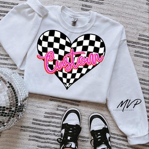 May include: White sweatshirt with a black and white checkered heart design and the word "Custom" in pink lettering. The sweatshirt has a crew neck and long sleeves.