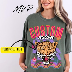 May include: Olive green t-shirt with a graphic design featuring a roaring leopard, the words "CUSTOM nation", and the text "YOUR MASCOT HERE". The shirt also has a star and paw print design.