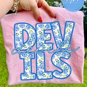 May include: A light pink t-shirt with the word "DEVILS" in large, blue-outlined letters filled with a blue rose pattern. The word "Devils" is written in a cursive font over the letters. The shirt is held up by a hand with red nail polish.