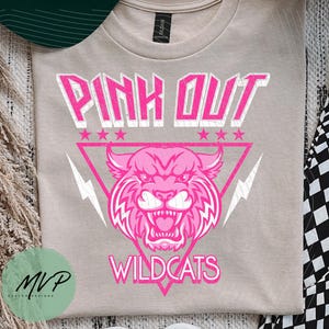 May include: A tan t-shirt with a pink graphic that reads "PINK OUT WILDCATS." The design features a pink wildcat head inside a pink triangle with lightning bolts and stars. A green and white baseball cap is visible in the upper left corner.