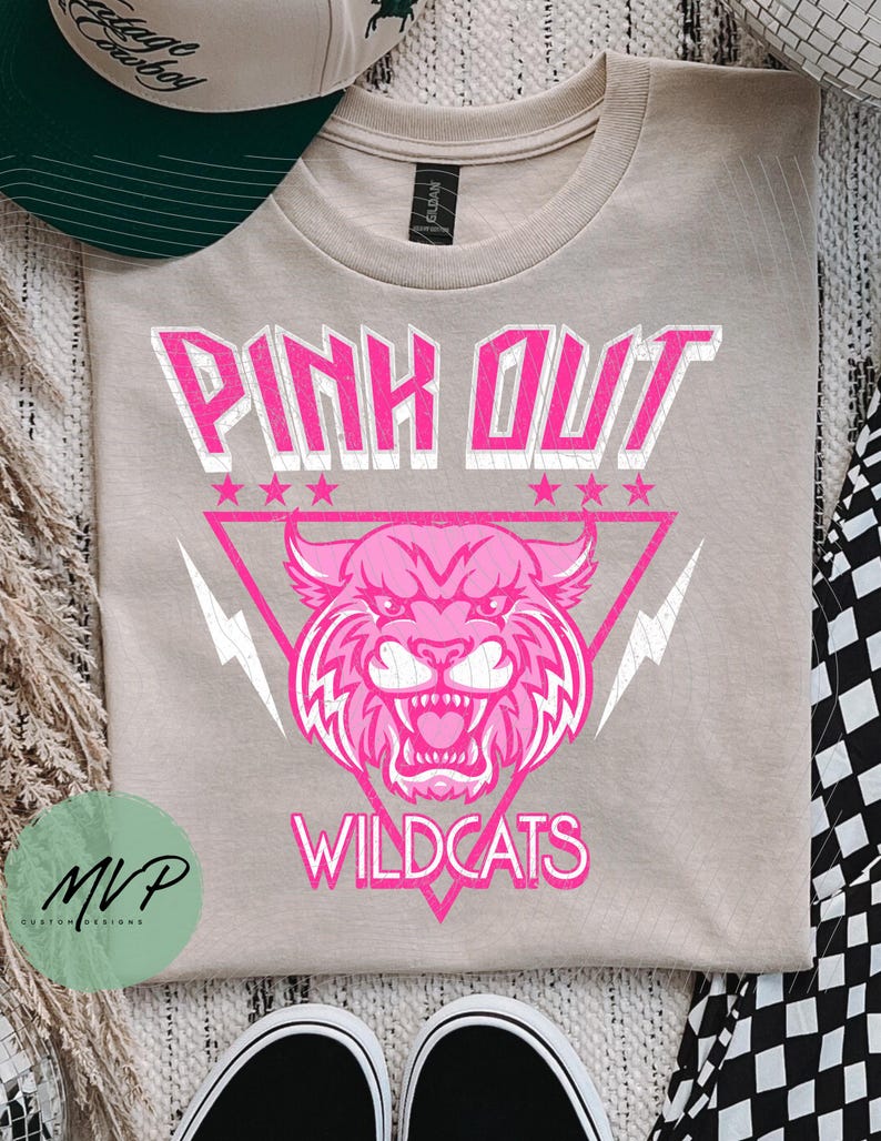 May include: A tan t-shirt with a pink graphic that reads "PINK OUT WILDCATS." The design features a pink wildcat head inside a pink triangle with lightning bolts and stars. A green and white baseball cap is visible in the upper left corner.
