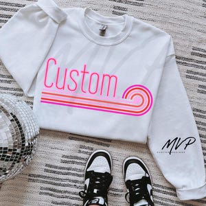 May include: White sweatshirt with the word "Custom" in pink neon lettering with a rainbow stripe design. The sweatshirt is on a white and grey patterned surface. The brand name "MVP Custom Designs" is visible on the bottom right corner of the sweatshirt.