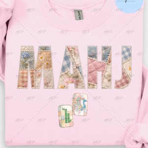 May include: A light pink sweatshirt with the word "MAHJ" in a patchwork design. The letters are made of various floral and patterned fabrics. Two small patches with a peacock and bamboo are below the letters.