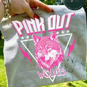 May include: A gray t-shirt with a pink wolf graphic and the words "PINK OUT WOLVES". The design includes a wolf's head, lightning bolts, and stars. The shirt is held up by a hand.