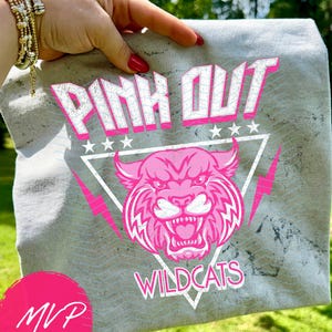 May include: A gray t-shirt with the words "PINK OUT" in white and pink lettering, along with a pink wildcat graphic. The shirt also features the word "WILDCATS" and a pink MVP logo.