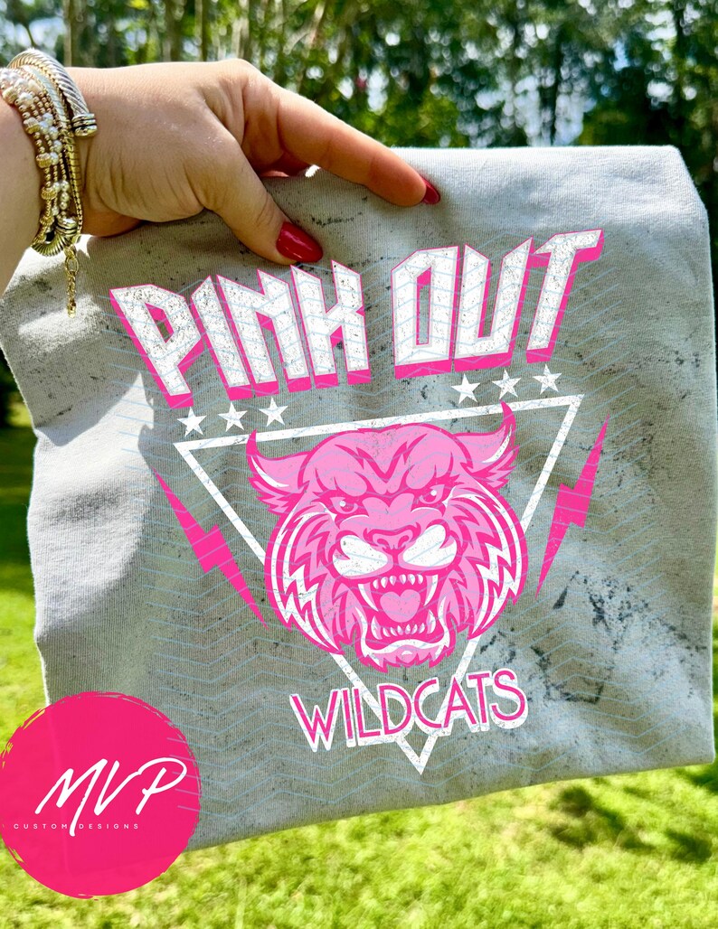 May include: A gray t-shirt with the words "PINK OUT" in white and pink lettering, along with a pink wildcat graphic. The shirt also features the word "WILDCATS" and a pink MVP logo.