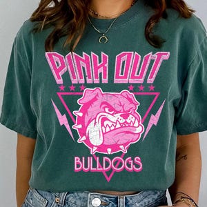 Może przedstawiać: Ciemnozielona koszulka z r&oacute;żową grafiką. Grafika przedstawia słowa "PINK OUT" nad r&oacute;żową głową buldoga w r&oacute;żowym tr&oacute;jkącie z błyskawicami i gwiazdami. Słowo "BULLDOGS" znajduje się poniżej.