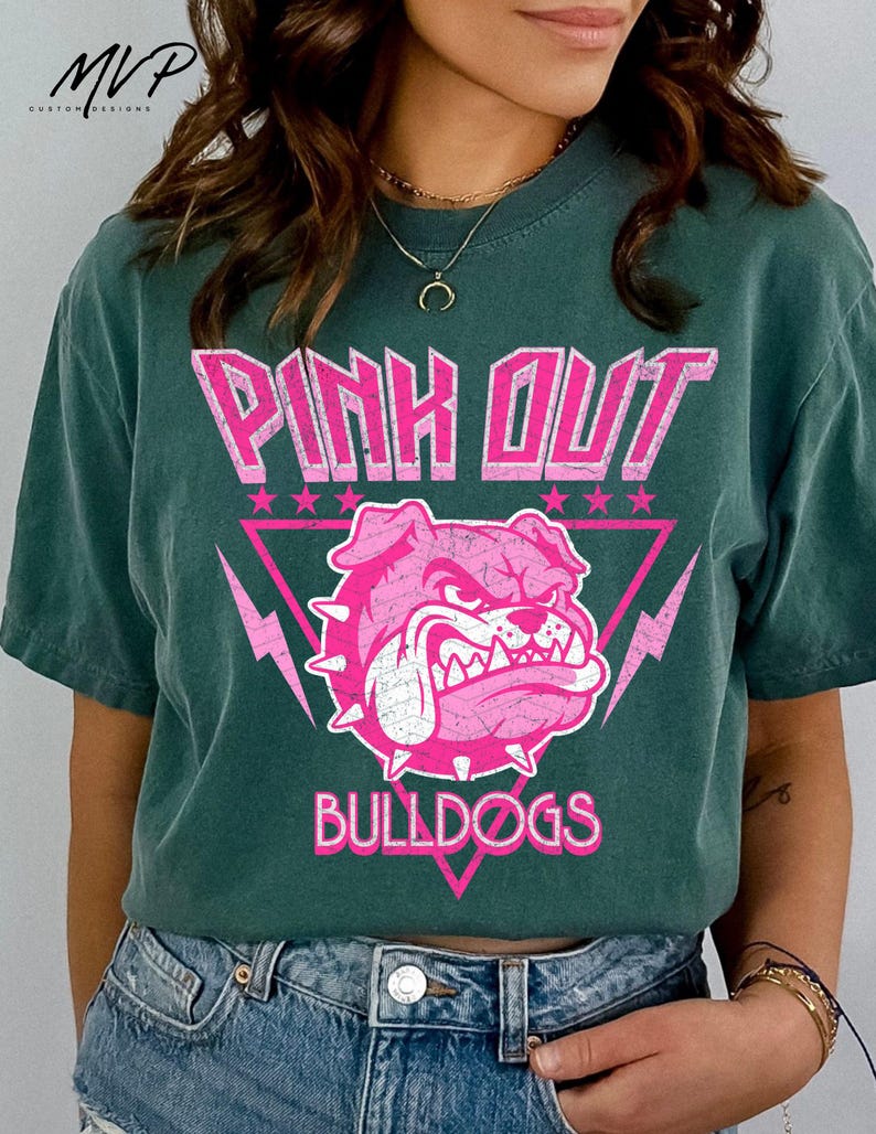 Może przedstawiać: Ciemnozielona koszulka z r&oacute;żową grafiką. Grafika przedstawia słowa "PINK OUT" nad r&oacute;żową głową buldoga w r&oacute;żowym tr&oacute;jkącie z błyskawicami i gwiazdami. Słowo "BULLDOGS" znajduje się poniżej.