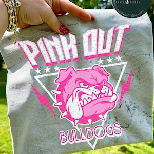Może przedstawiać: Szara koszulka z napisem "PINK OUT" w kolorze białym i r&oacute;żowym, r&oacute;żową grafiką buldoga i słowem "BULLDOGS". Projekt zawiera gwiazdy i błyskawice.