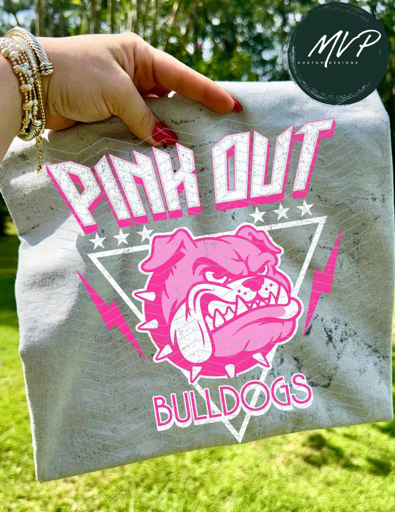 Może przedstawiać: Szara koszulka z napisem "PINK OUT" w kolorze białym i r&oacute;żowym, r&oacute;żową grafiką buldoga i słowem "BULLDOGS". Projekt zawiera gwiazdy i błyskawice.