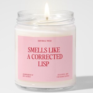 May include: A lit candle in a clear glass jar with a pink label. The label reads "Smells Like a Corrected Lisp" in red text. The candle is on a white background.