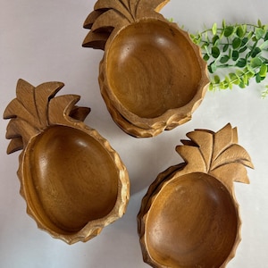 May include: Three wooden pineapple-shaped bowls. The bowls are carved from light brown wood, with detailed leaf-like tops. The bowls are arranged on a white surface, suggesting a tropical theme. The bowls are likely for serving snacks or displaying decorative items.