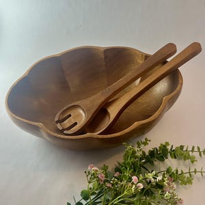 May include: A wooden salad bowl with a scalloped edge, accompanied by two wooden salad servers. The bowl and servers are made of a warm-toned wood. A sprig of greenery with small pink flowers is in the foreground.