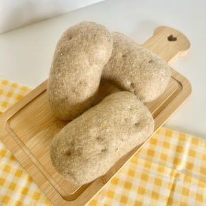 May include: Three beige felt potatoes rest on a small wooden cutting board with a heart-shaped cutout. The potatoes have a textured appearance and are arranged on a yellow and white checkered cloth.